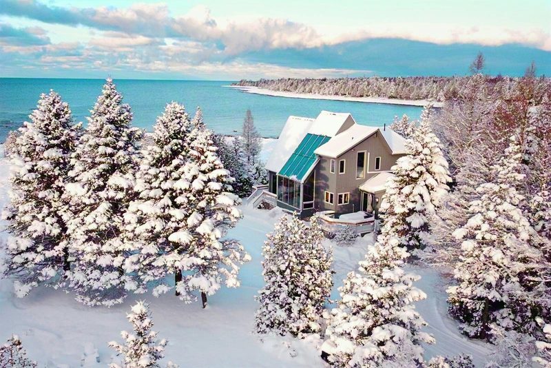 Casolena from above after heavy snow: the house among snow-laden evergreens, with the open turquoise water of Lake Huron and a white shoreline curving away behind it