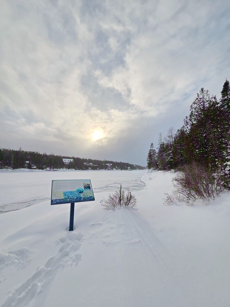 A Georgian Bay Coastal Route interpretive sign standing in deep unbroken snow with the low winter sun behind cloud