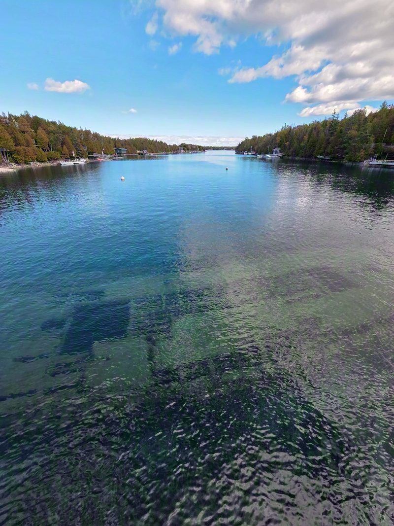Big Tub Harbour on a calm day with the dark shape of a wreck showing through clear water and boathouses along the shore