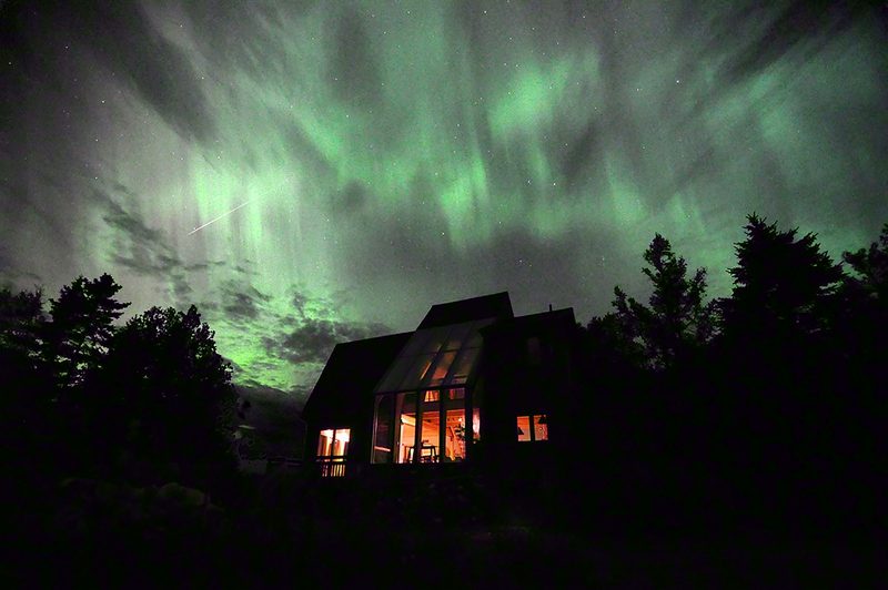Green northern lights spread across the whole sky above the cottage, with a meteor streak to the left