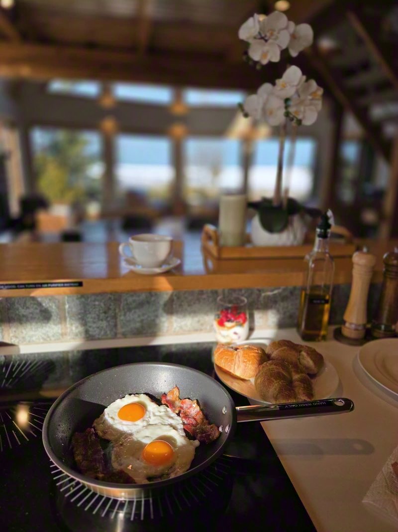 Eggs and bacon in a pan on the induction cooktop, croissants and coffee beside it and the great room windows behind