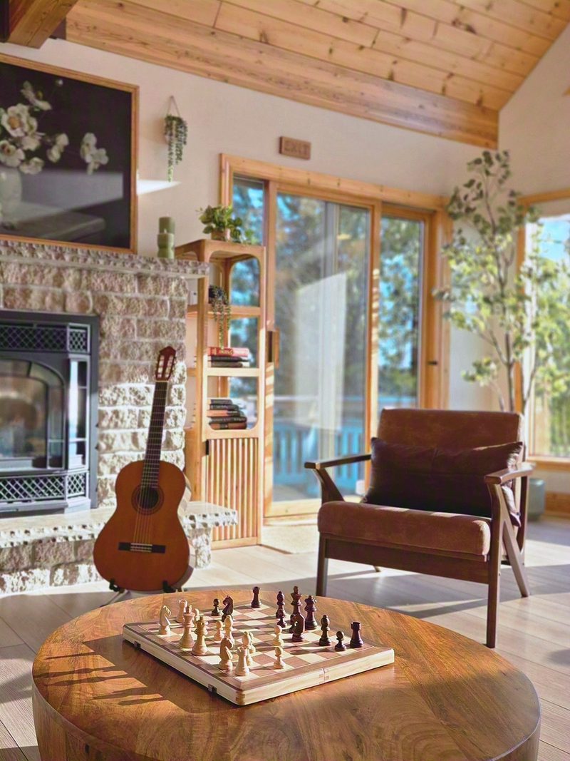 A cottage great room with a stone fireplace, a guitar, a chess board on a round wood table and sliding doors opening to the trees and the water