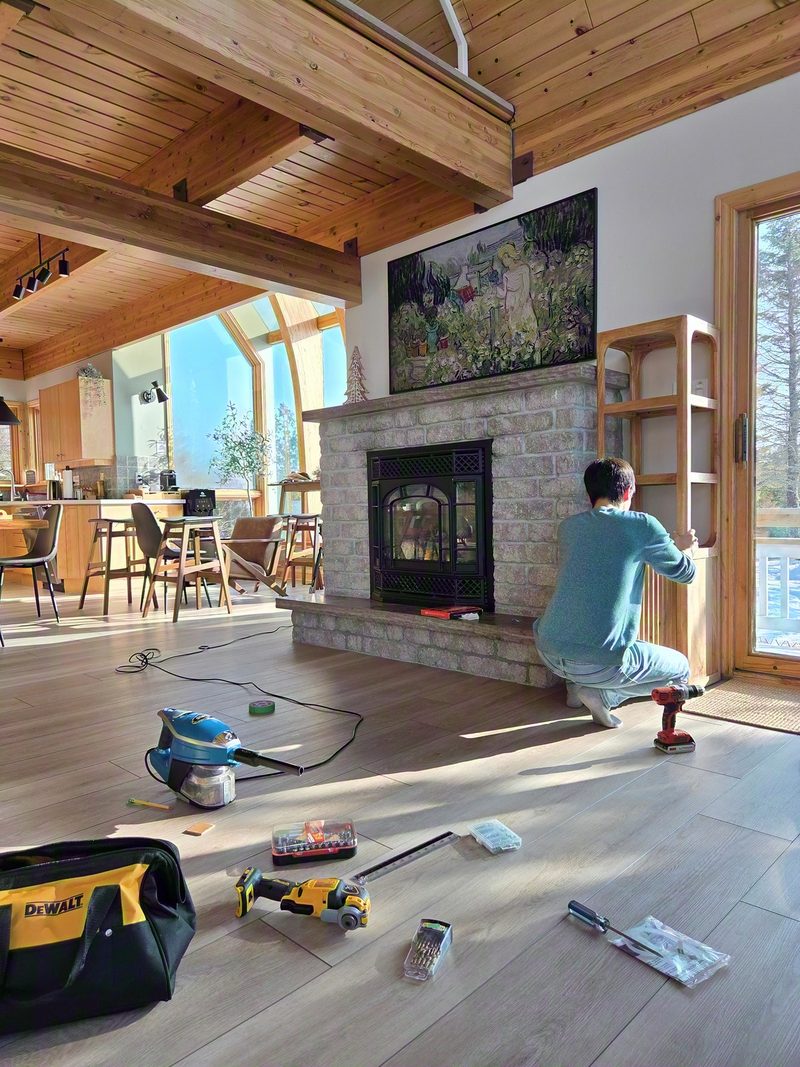 Hoan kneeling with a cordless drill, assembling a wooden shelf frame by the window beside the stone fireplace, tools spread across the floor