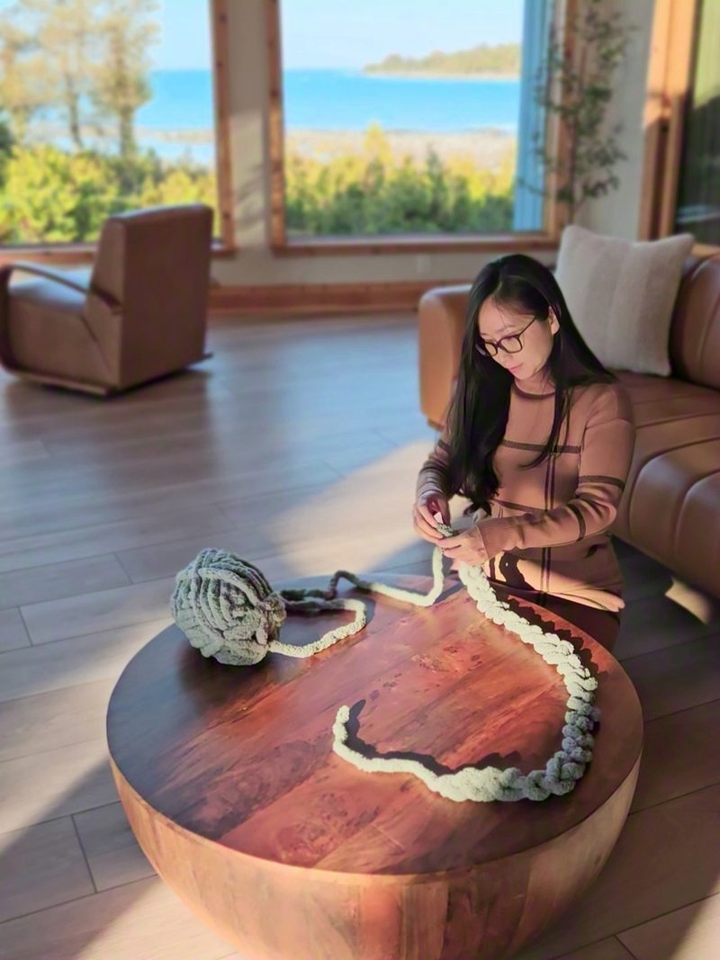 Jenny kneeling at the round wood coffee table working a chunky knit blanket, the lake through the windows behind her