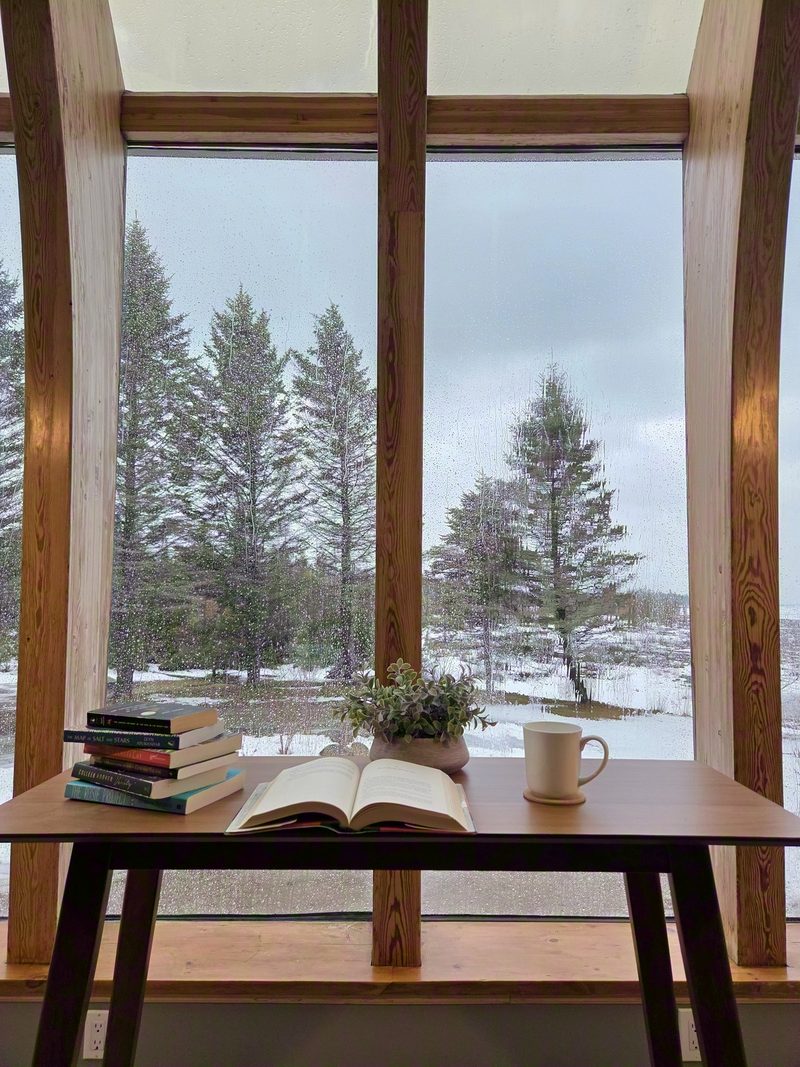 An open book and a mug on a bay window table, rain on the glass and snowy trees outside
