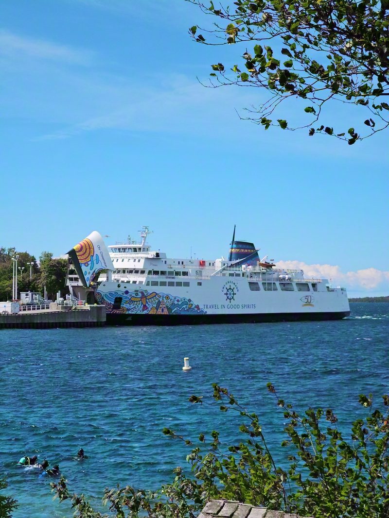 The MS Chi-Cheemaun at her Tobermory berth with the bow door raised, the painted hull and TRAVEL IN GOOD SPIRITS along the side, seen from the shore through branches