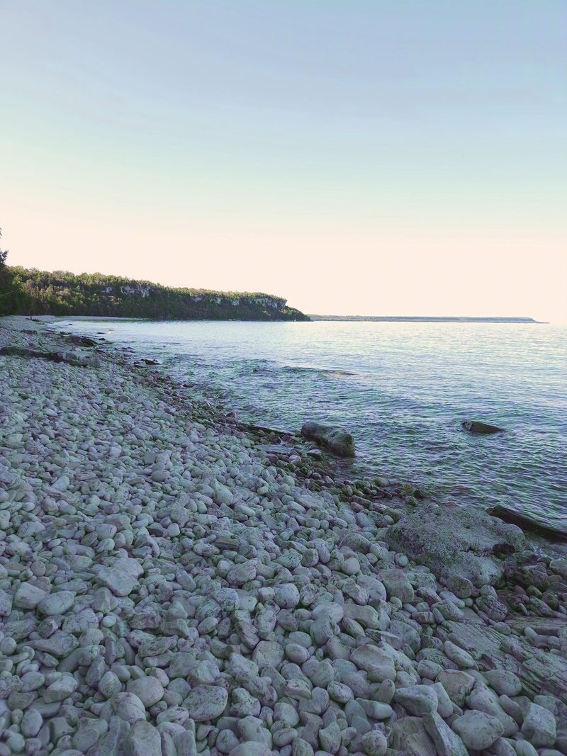 A white cobble beach in afternoon shade, curving toward a sunlit forested headland