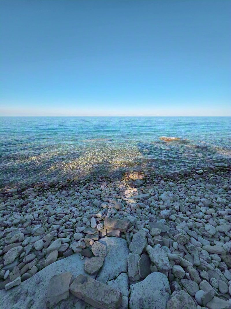 The cobble beach and clear blue water below Devil's Monument, mostly in shade with one patch of sun reaching the shore