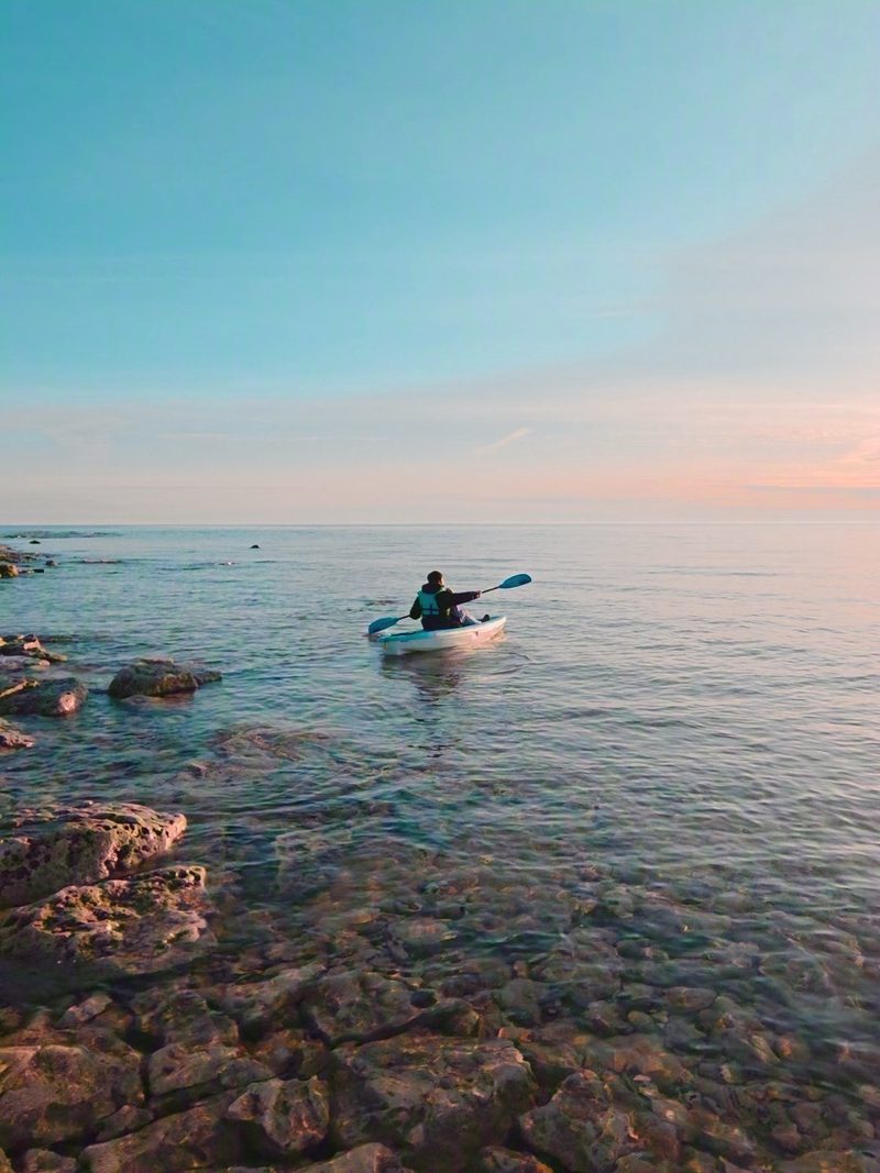 A kayaker in a PFD paddling calm water along a rocky shore under a soft dusk sky