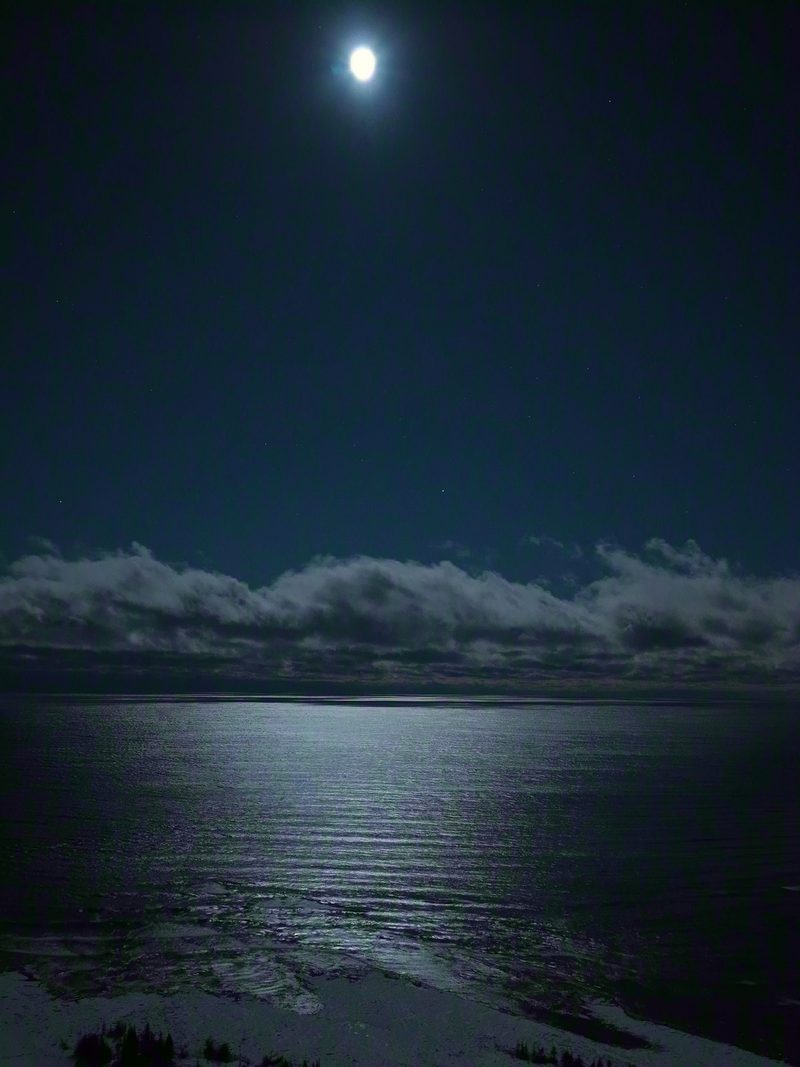 A full moon high over Lake Huron just after midnight, laying a broad path of light across the water to a cloud bank
