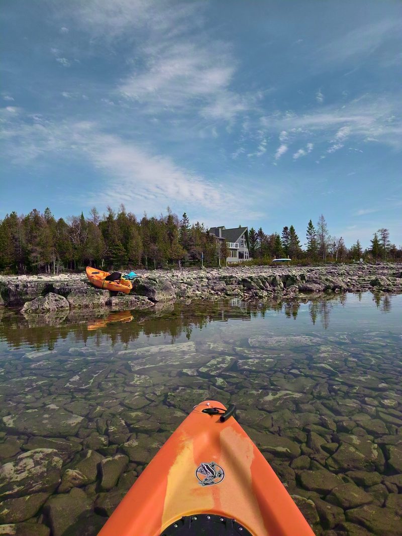 The bow of a kayak over clear water and a boulder bottom, with more kayaks waiting on the rocky shore ridge behind
