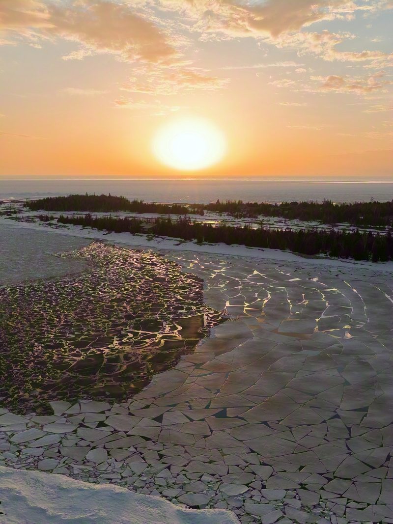 Golden sunset over a shore of broken ice plates and open water, seen from above with a forested headland