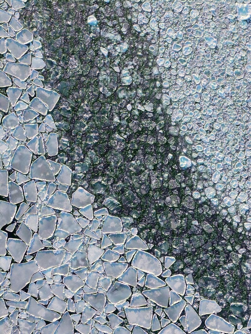 Broken ice plates over dark water seen straight down from above, a mosaic of white and grey shards