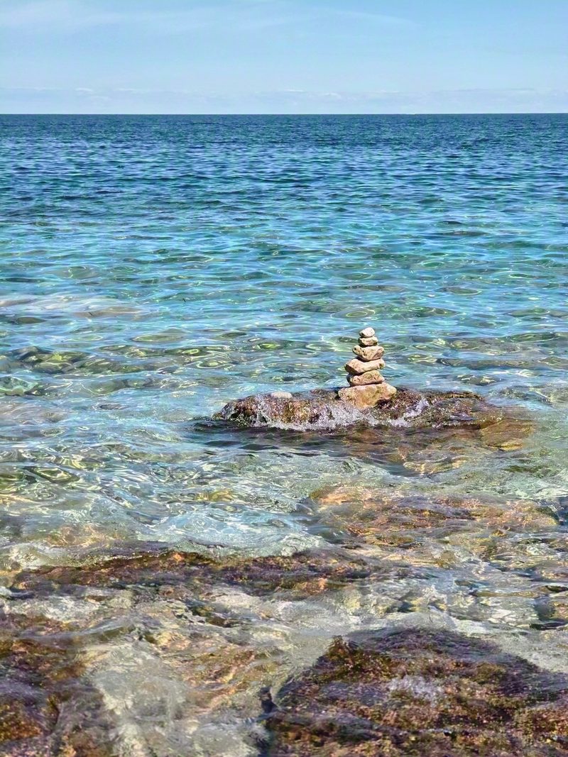 A small stone cairn balanced on a rock in the clear shallows at Little Cove with open water behind