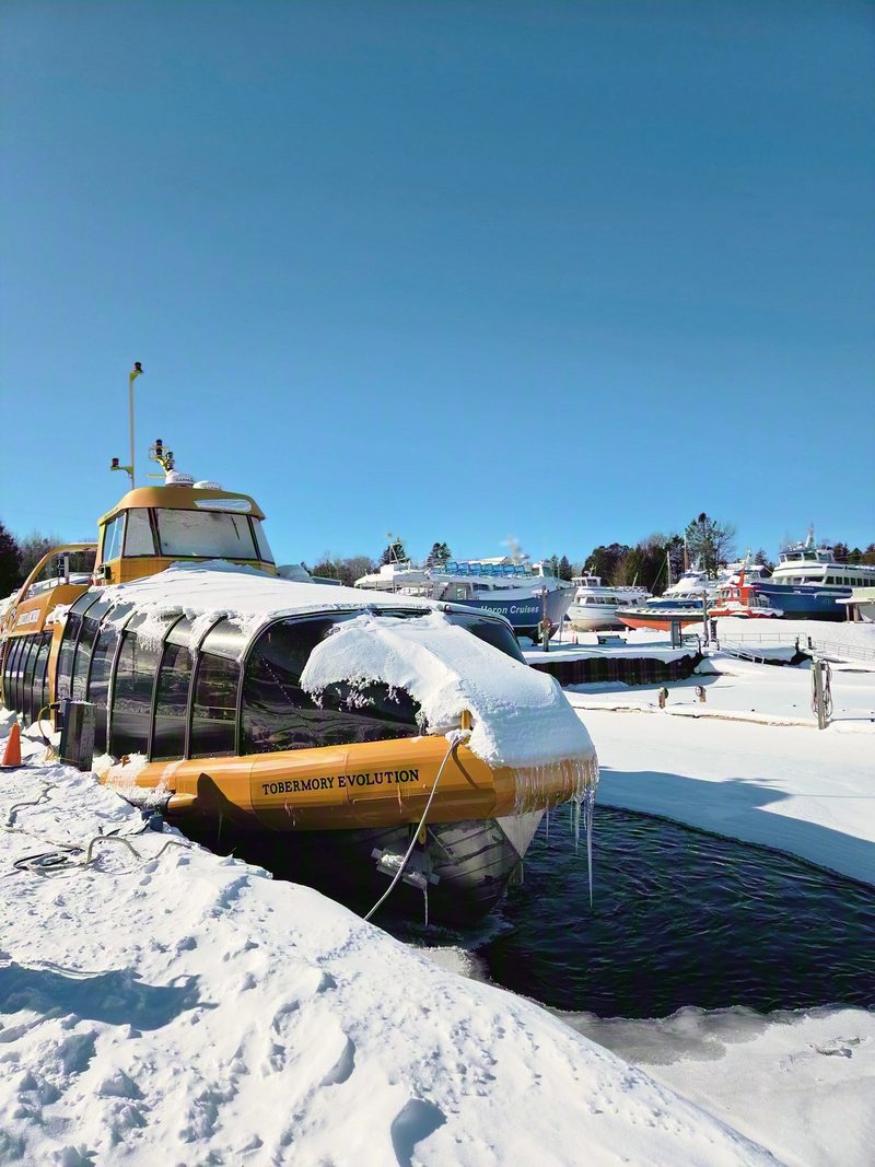 A yellow glass-bottom boat on blocks in the snow with dark open water of the harbour behind it