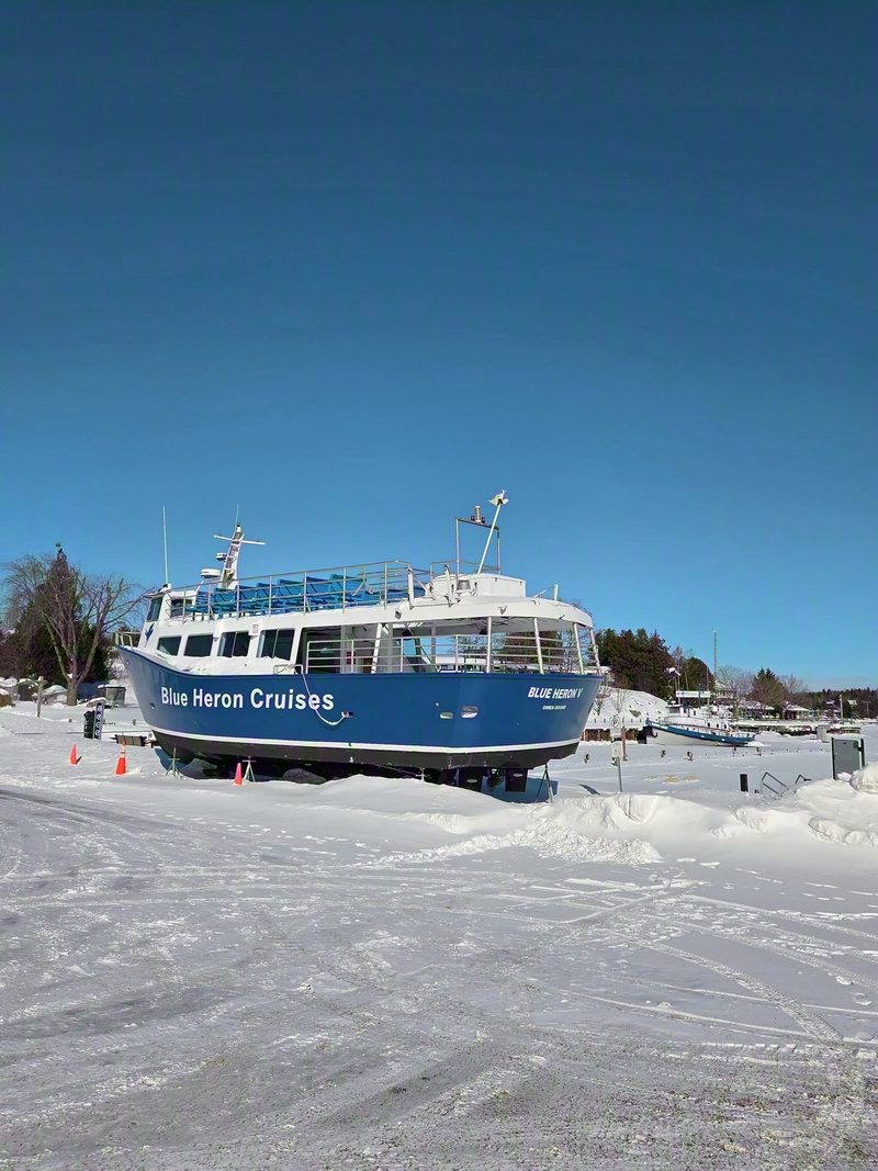 The Blue Heron V tour boat up on blocks in a snow-covered parking lot at Little Tub Harbour under a clear blue sky