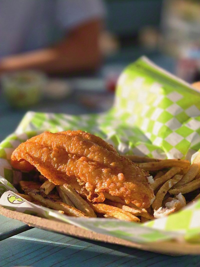 Battered fish and fries in a green checkered basket on a blue picnic table in evening light