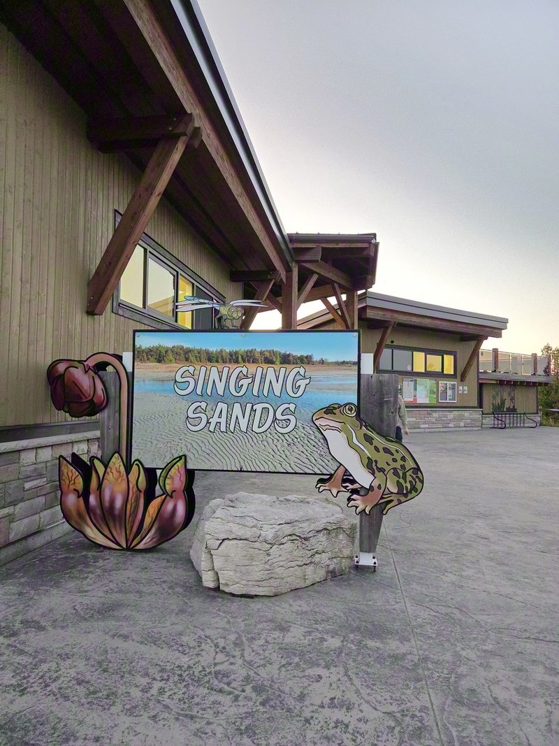 The Singing Sands entrance sign with cutout frog and orchid illustrations beside the visitor building