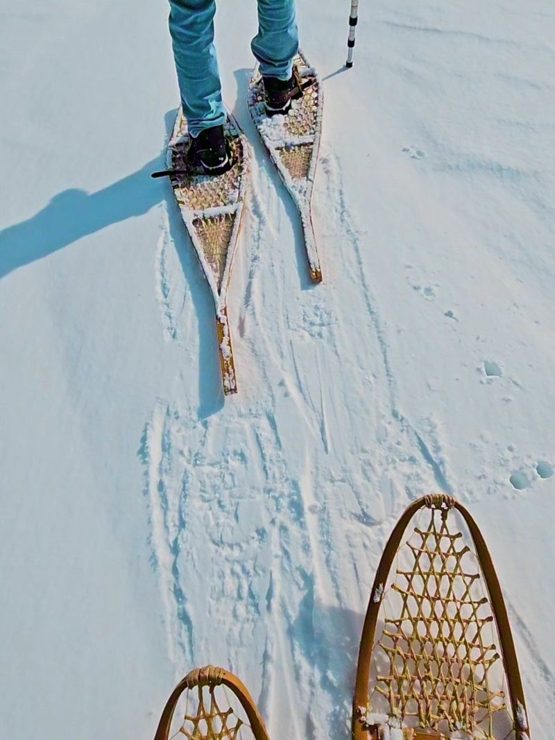 Looking down at traditional wooden snowshoes mid-stride on packed snow, a second pair carried ahead