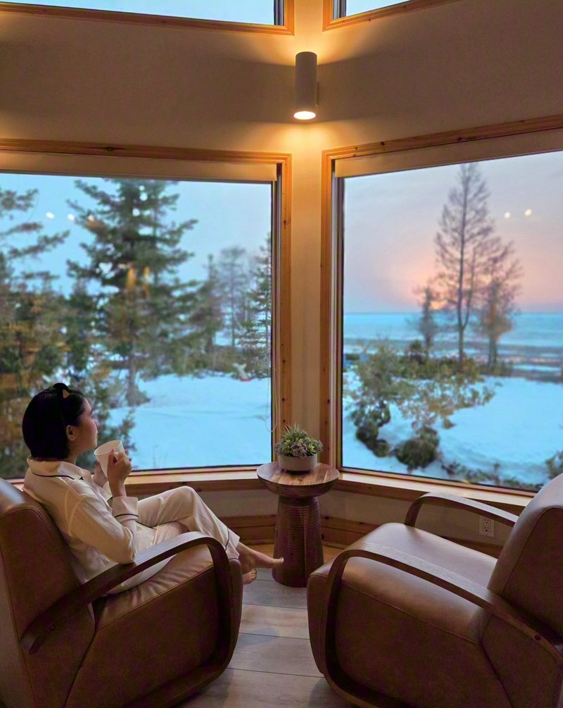 A guest with a mug in the leather armchair by the corner windows as the sun sets over the lake
