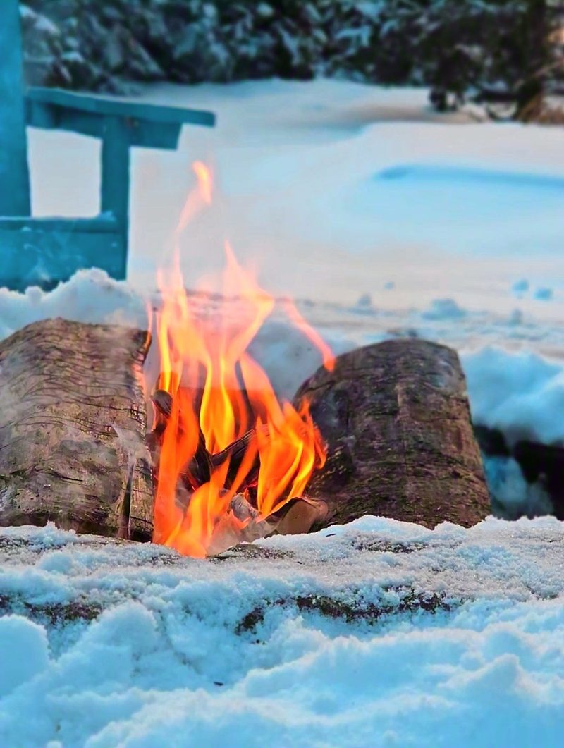 A fire burning between two logs in deep snow with a blue Adirondack chair and spruces behind