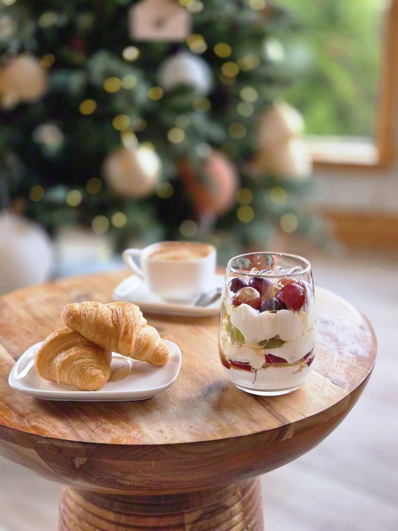 Croissants, a latte and a berry parfait on a round wooden table with a lit Christmas tree soft behind