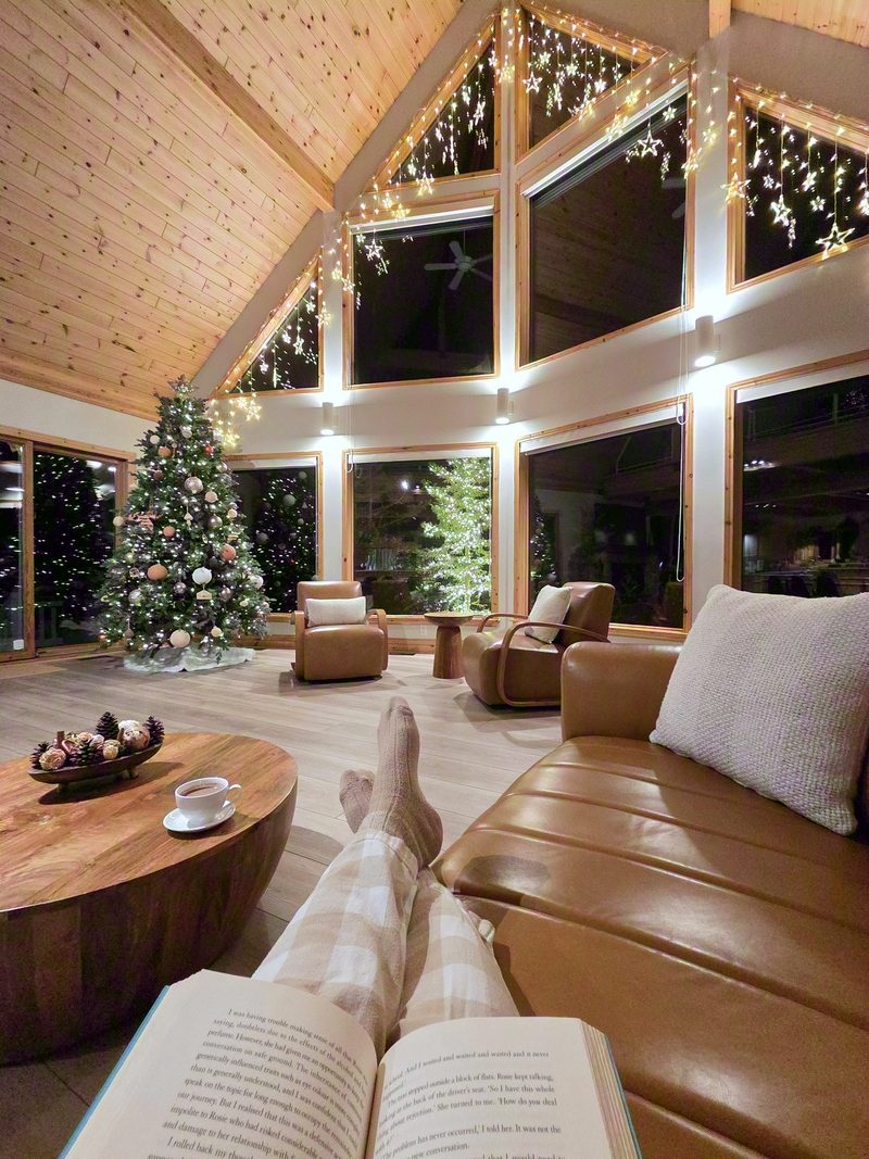 Reader's view over an open book and stockinged feet on the leather couch, two lit Christmas trees beyond