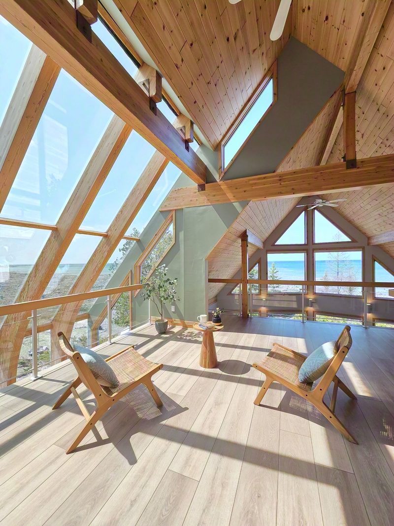 The Cielo loft at Casolena: angled lake and sky windows following the roofline, two rattan chairs and Lake Huron beyond the glass