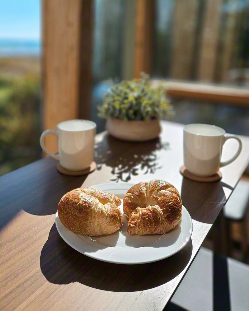 Two croissants, two mugs and a small potted plant on the table in hard morning light, the lake beyond