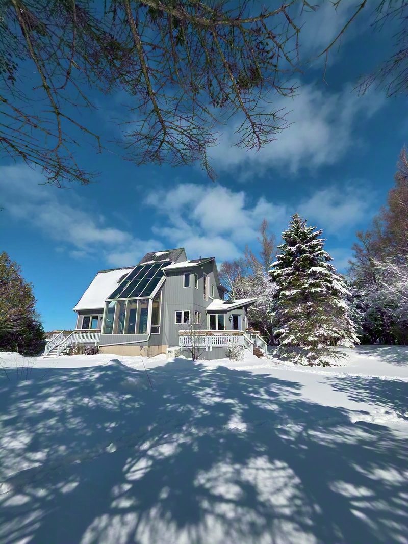 The cottage and its tall sunroom glazing surrounded by deep snow and snow-laden evergreens