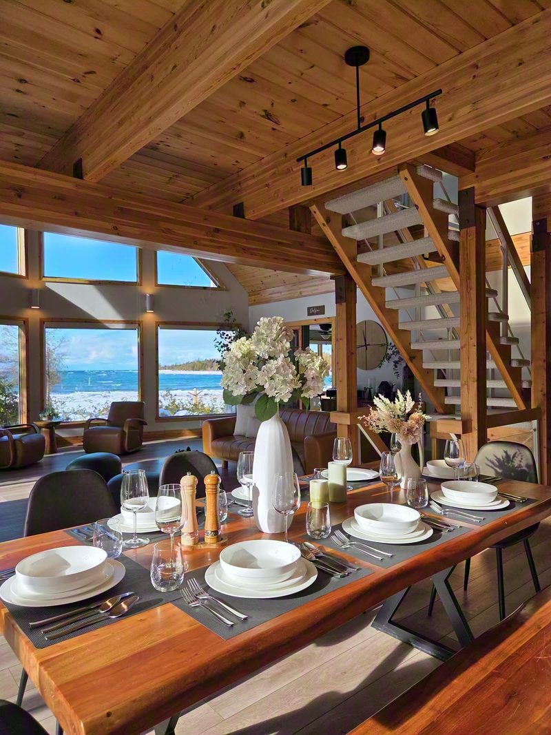 Live-edge dining table set with white dishes and flowers, staircase and lake windows behind