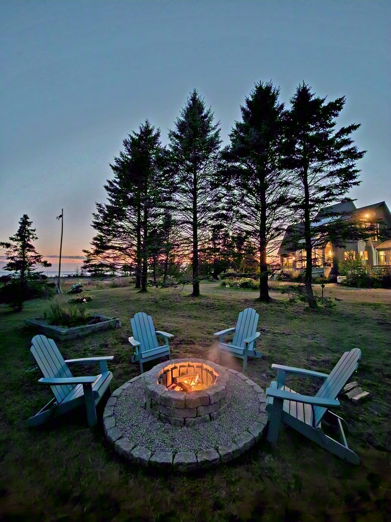 Stone fire pit burning at dusk ringed by Adirondack chairs, tall spruces and a sunset sky