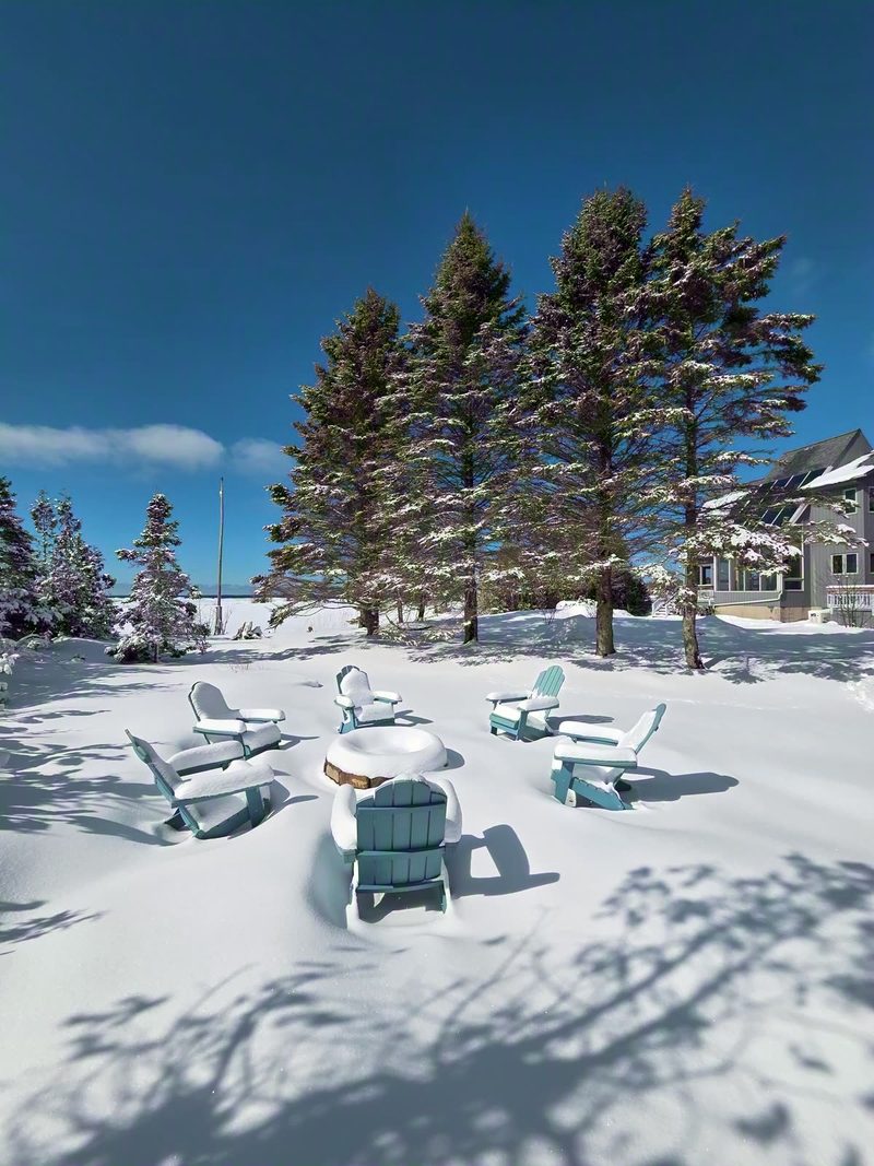 Fire pit and Adirondack chairs buried in deep snow with snow-covered spruces behind
