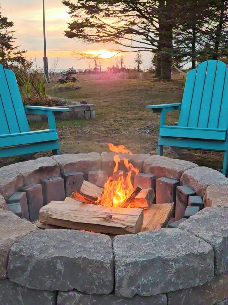 A small fire burning in a stone fire pit ring on cleared ground, with 2 teal chairs and the sunset over the lake behind