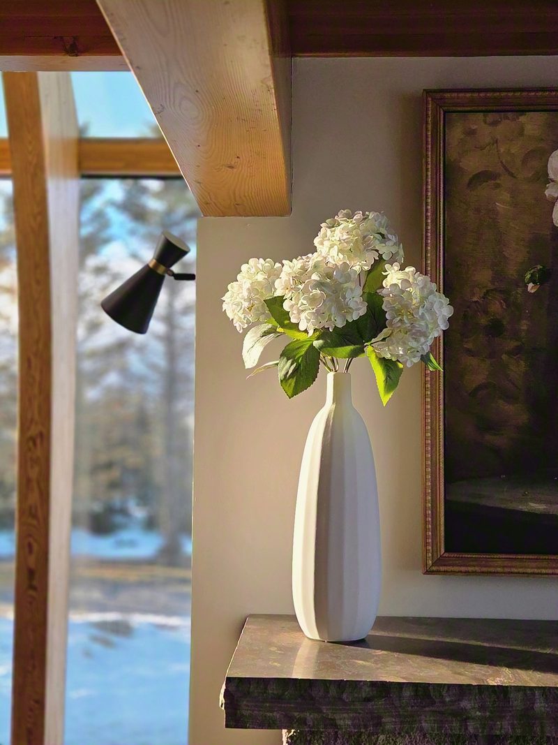 White hydrangeas in a tall ceramic vase on a wooden ledge beside a gilt-framed painting