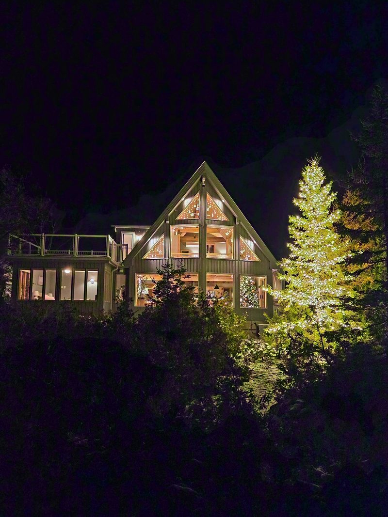 The A-frame lit from within on a dark winter night with a decorated spruce glowing beside it