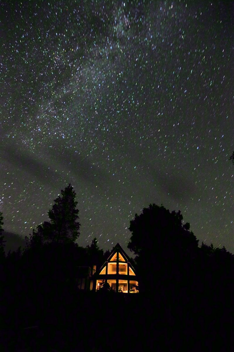 The Milky Way over dark trees and the warmly glowing A-frame windows of the cottage at night