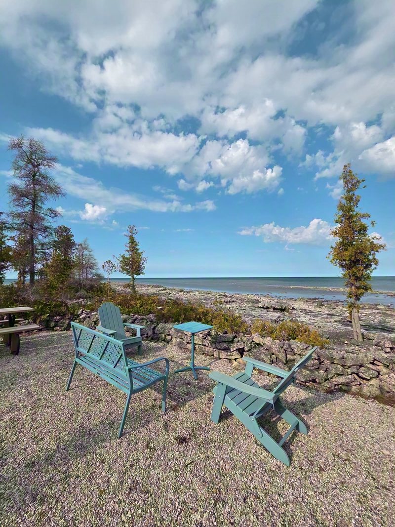 Teal Adirondack chairs and side table on gravel at the water's edge under a jet-trail sky