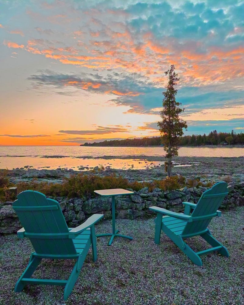 Two teal Adirondack chairs on the shore under a peach and blue sunset sky