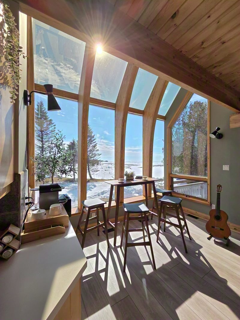 A bar table with stools and a kettle beside the two-story glass wall, guitar waiting, snowy trees outside