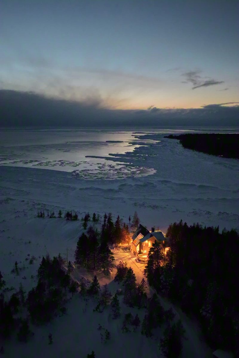 Aerial view at dusk of the warmly glowing cottage in deep snow beside the icy lake