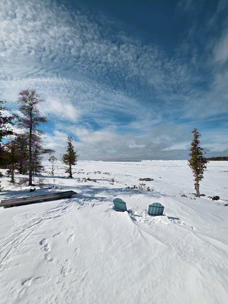 Two Adirondack chairs on the snow-covered shoreline facing the frozen lake under a dramatic sky