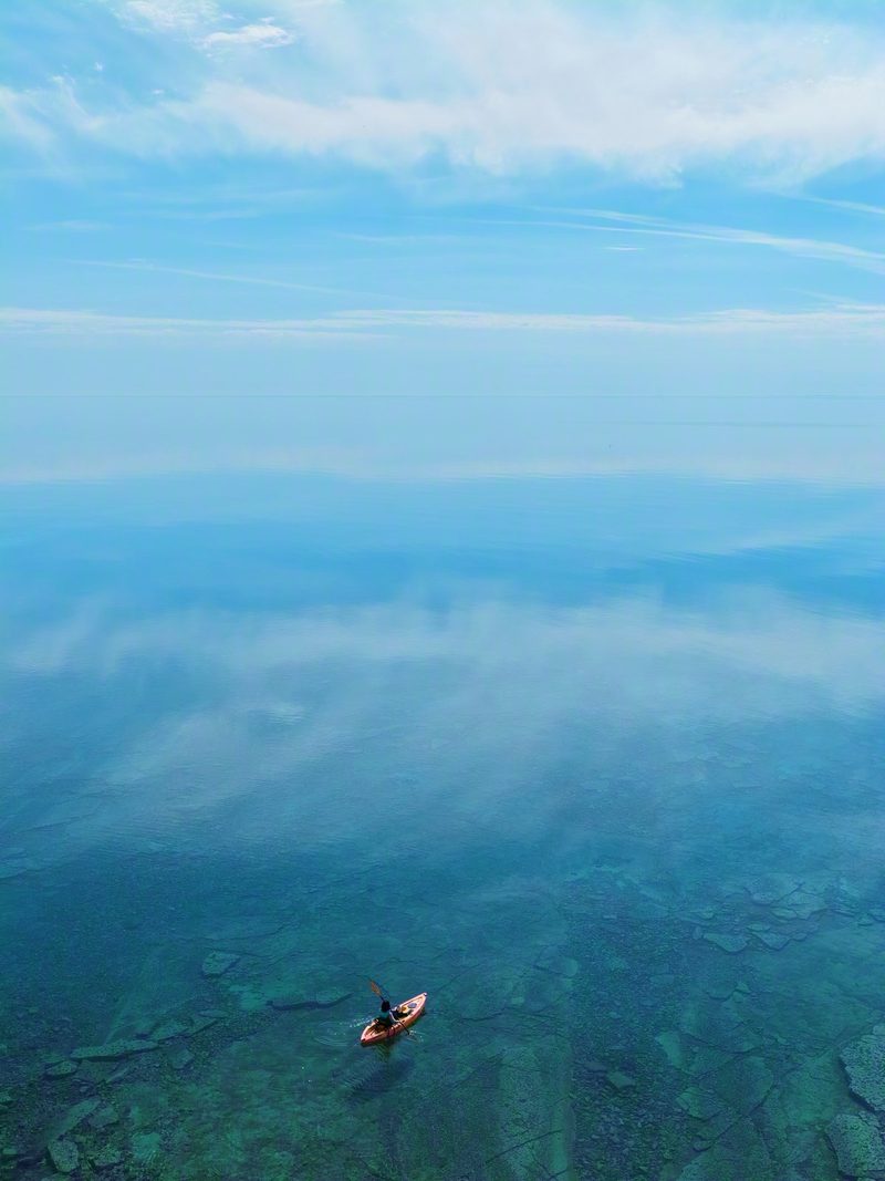A lone kayaker on glassy Lake Huron from above, clear water fading from turquoise over limestone to open blue