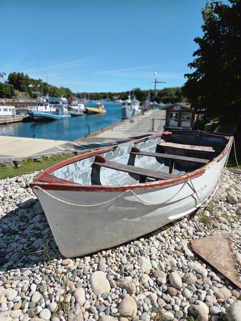 An old white and red wooden rowboat resting on the cobble above Little Tub Harbour with the tour boats behind